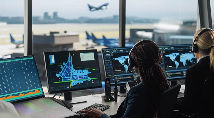 Two air traffic controllers wearing headsets monitor multiple screens displaying flight data and radar maps in a control
