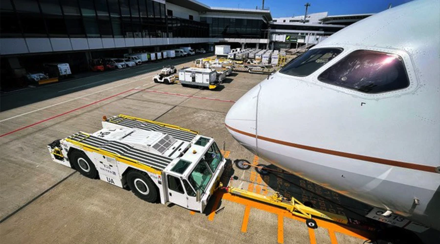 Airport ground crew tug vehicle positioned at aircraft nose on tarmac with terminal and cargo equipment in background