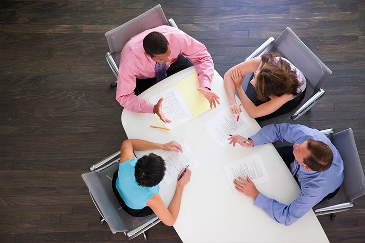Four business professionals in meeting reviewing documents around white oval conference table viewed from above